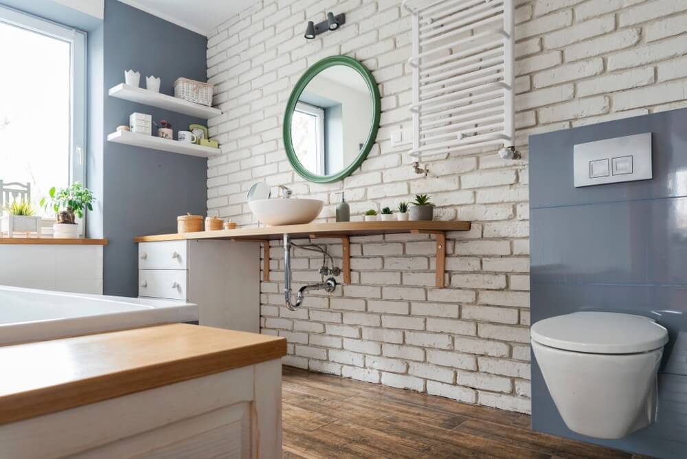 Interior of bathroom in white and grey color with wooden furniture, round mirror and brick wall. 